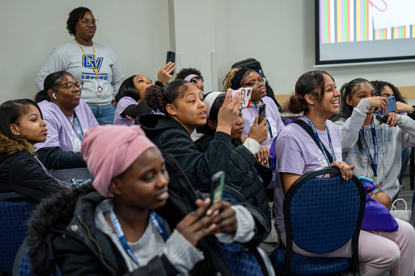 A group of young people in a conference room are seated, many holding phones, while one person stands in the background.